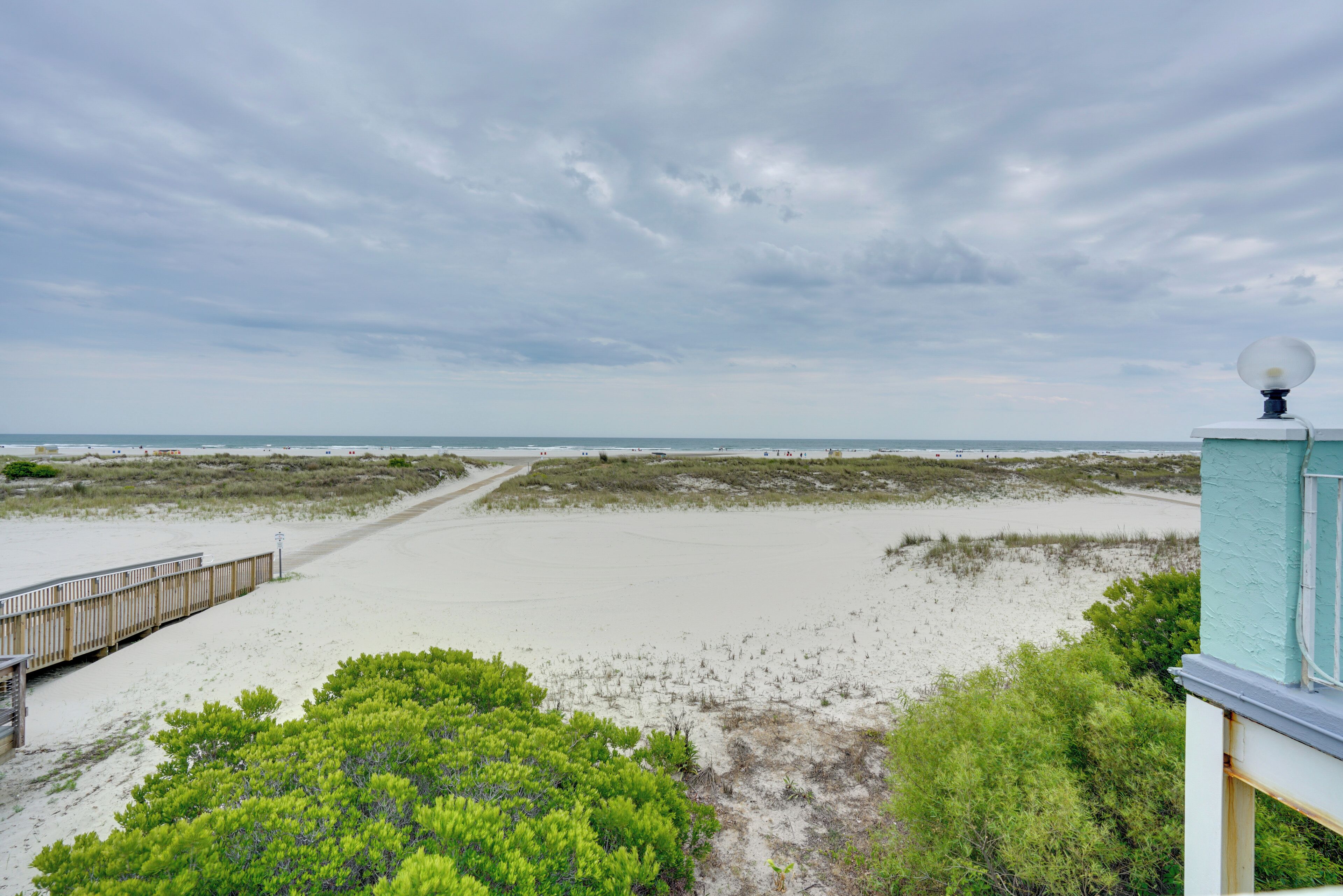 Vista de una playa con un paseo marítimo de madera que conduce al agua, rodeado de vegetación verde.