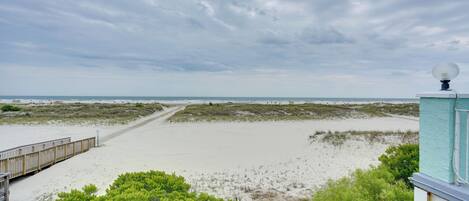Vista de una playa con un paseo marítimo de madera que conduce al agua, rodeado de vegetación verde.