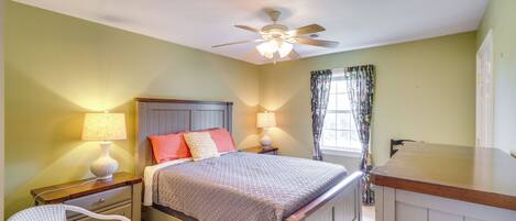 A bedroom with a bed, two bedside tables with lamps, a chair, a ceiling fan, and a window with curtains.