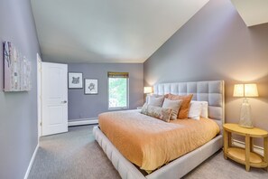 Ein Schlafzimmer mit Bett, Nachttisch, Lampe, einer Tür und gerahmten Bildern an der Wand.