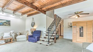 A spacious living area with a high ceiling, a wooden staircase, a blue armchair, a beige sofa, and a wooden door.