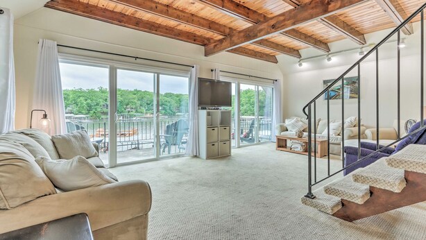A spacious living room with a high ceiling, large windows, a staircase, and a flat-screen TV.