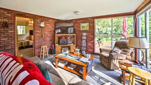 A cozy living room with a brick wall, a television, a bookshelf, and a large window with a view of greenery.