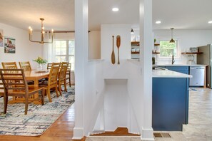 Une cuisine moderne avec des meubles blancs, un îlot de couleur bleue, des chaises en bois disposées autour d’une table à manger, et un mur décoratif orné de deux pagaies en bois.