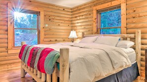 A wooden cabin bedroom with a bed, a nightstand, and two windows. - Cedar Mountain Log Cabin: 4 Mi Dupont State Forest (Brevard)