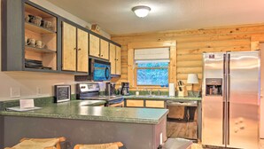 A kitchen with wooden cabinets, a stainless steel refrigerator, a microwave, and a coffee maker. There are two wooden stools and a window with a view of trees. - Cedar Mountain Log Cabin: 4 Mi Dupont State Forest (Brevard)