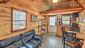 A cabin interior with a wooden interior, a leather couch, a dining table with chairs, a kitchen area, and a loft bedroom with a wooden railing. - 'the Gosling' Studio Cabin w/ River Access! (New Bern)