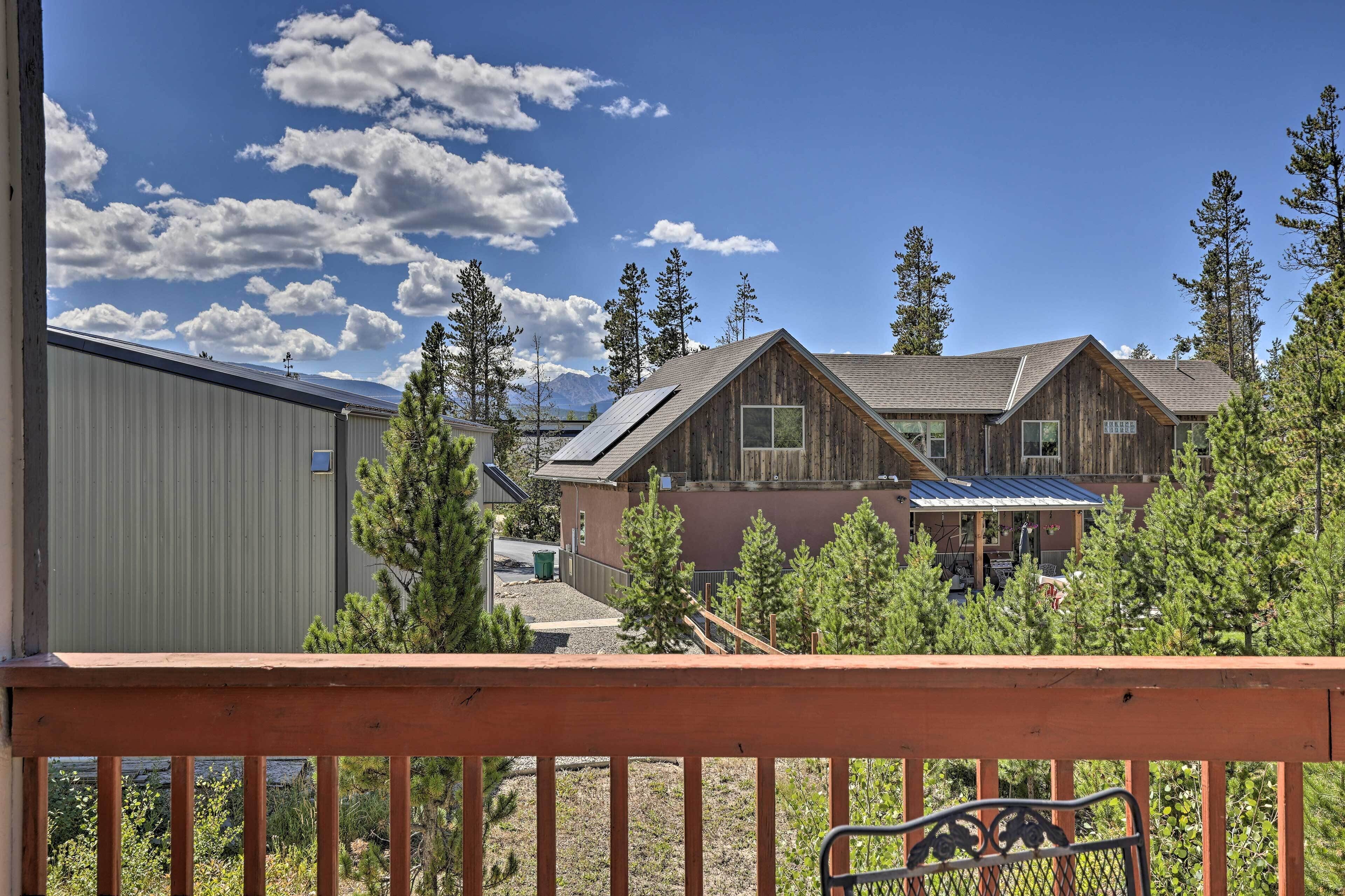 A wooden cabin with a large deck, surrounded by trees and a mountain in the background.