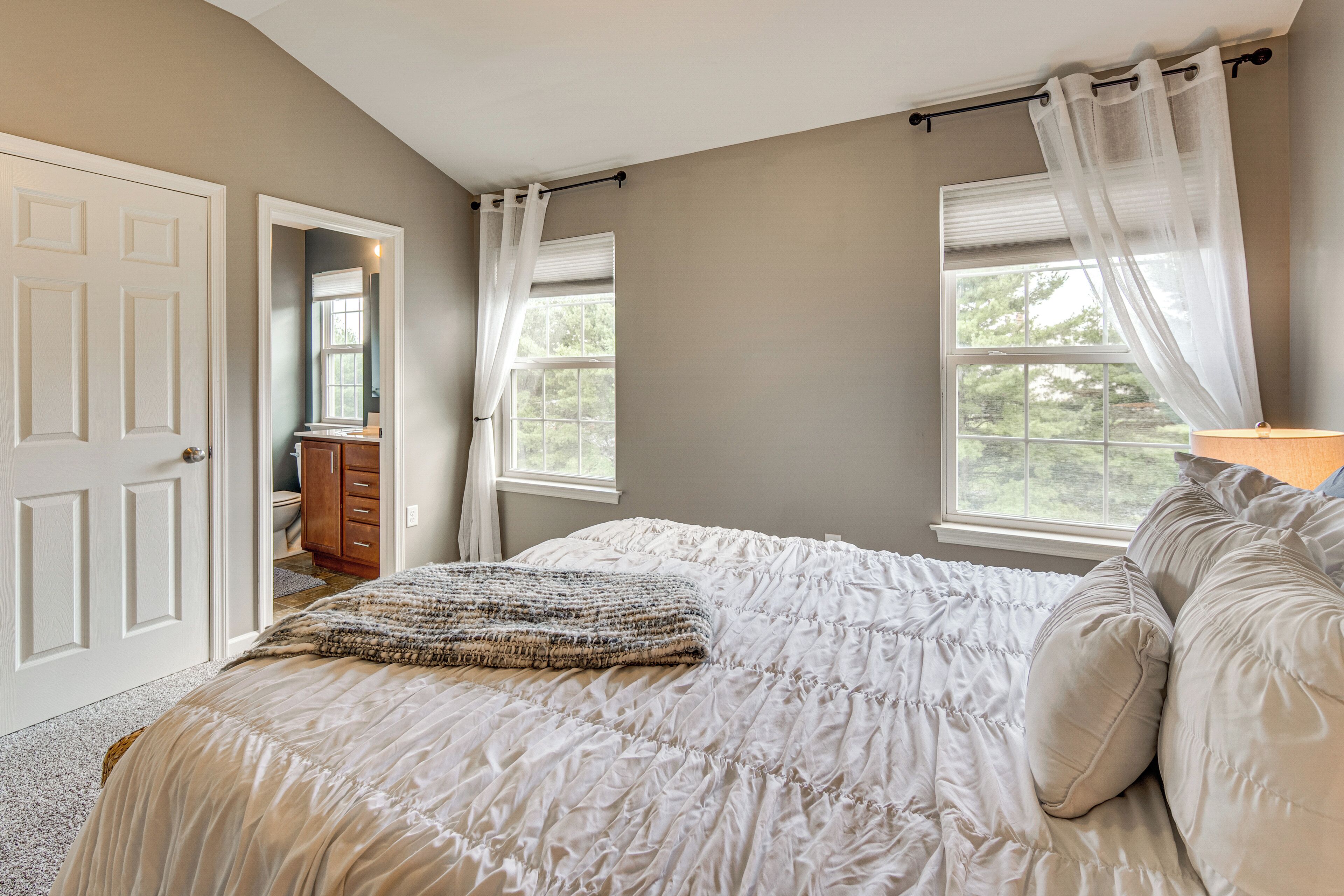 A bedroom with a large bed, two windows with curtains, a door, and a dresser.