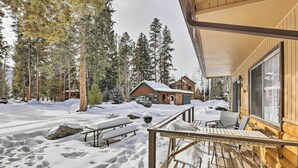 A wooden cabin with a deck, surrounded by snow-covered ground and tall pine trees.