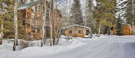 Une cabane en bois recouverte de neige, dotée d’un porche et entourée de grands pins.