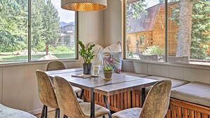 A dining area with a wooden table and chairs, a bench with cushions, and a view of a wooden cabin through large windows.