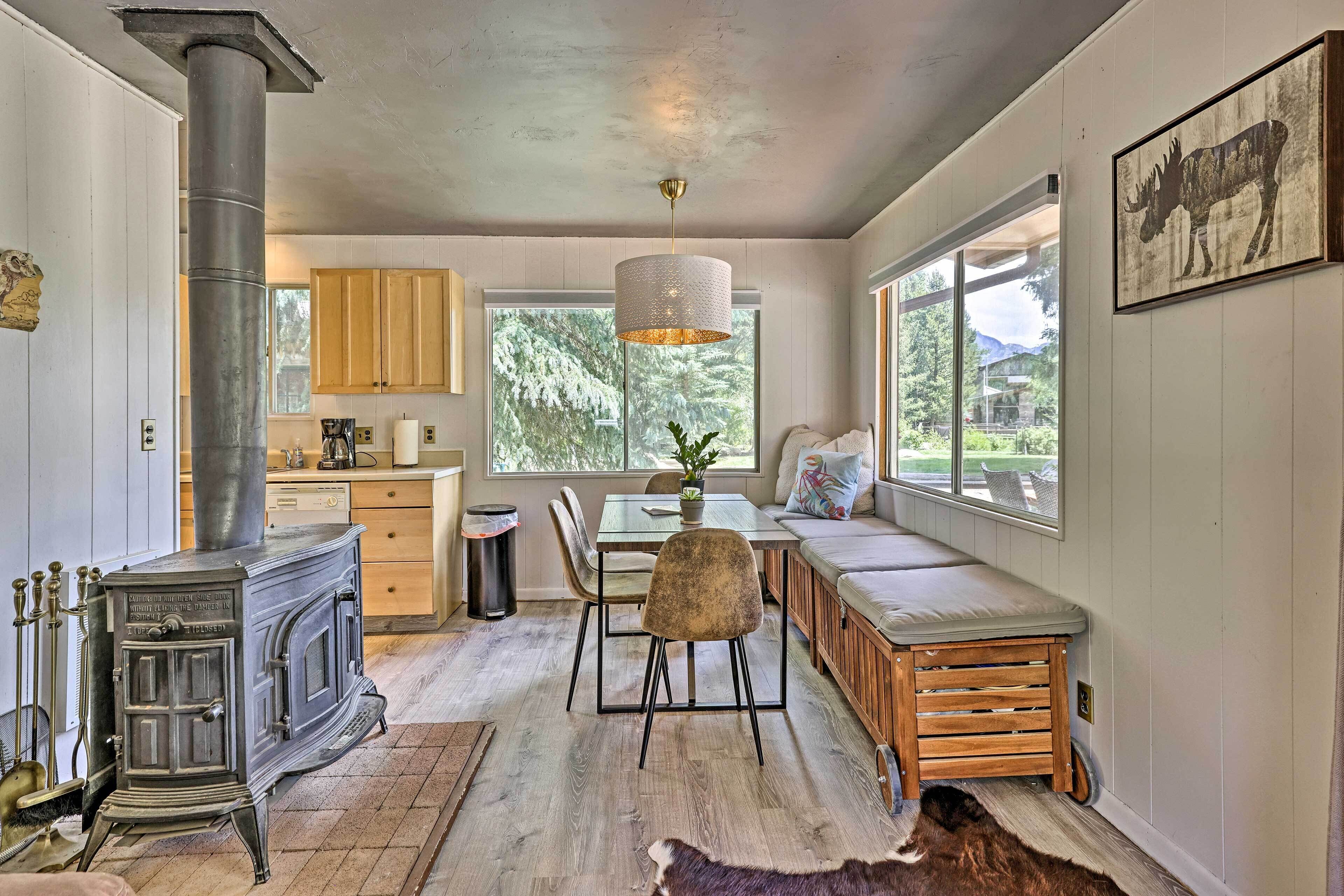 A cozy interior with a wood-burning stove, a dining area, and a large window offering a view of the outdoors.