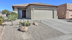 Une maison de plain-pied avec un garage, entourée d’une allée en gravier et d’un aménagement paysager de type désertique comprenant des cactus et de petits arbres.