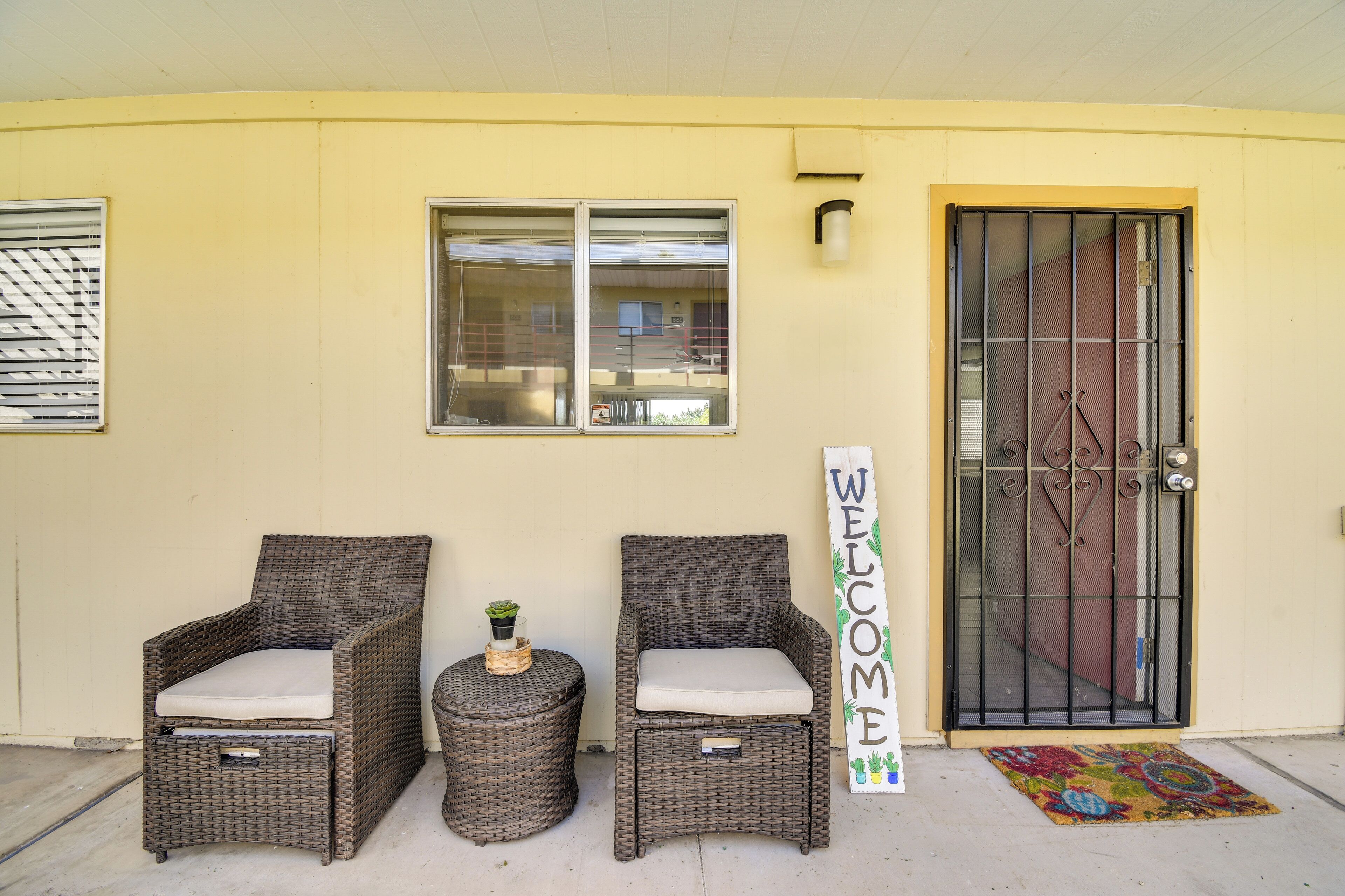 Two wicker chairs with cushions, a small round table with a plant, and a 'WELCOME' sign beside a door with a metal gate.