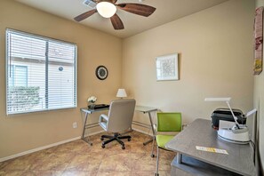 Ein Zimmer mit Deckenventilator, Schreibtisch mit Drucker, Stuhl, Lampe und Fenster mit Jalousien.