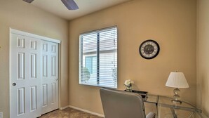 Una habitación con una puerta blanca, una ventana con persianas, un reloj, una mesa de vidrio con una lámpara y una silla verde.