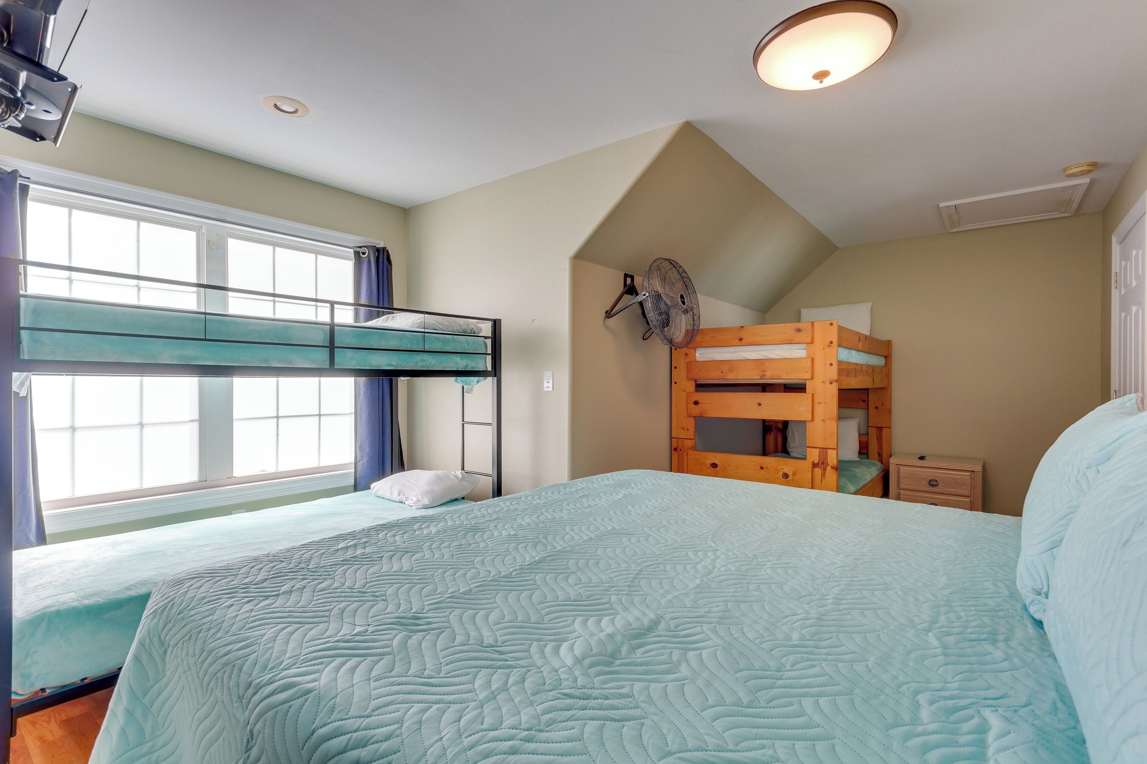 A dormitory room with bunk beds, a large bed, a ceiling fan, and a window with curtains.