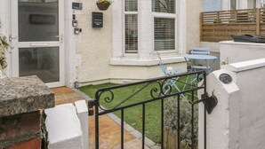 Una propiedad residencial con una puerta blanca, una ventana con contraventanas, un pequeño jardín con un portón de metal y una silla azul en el exterior.