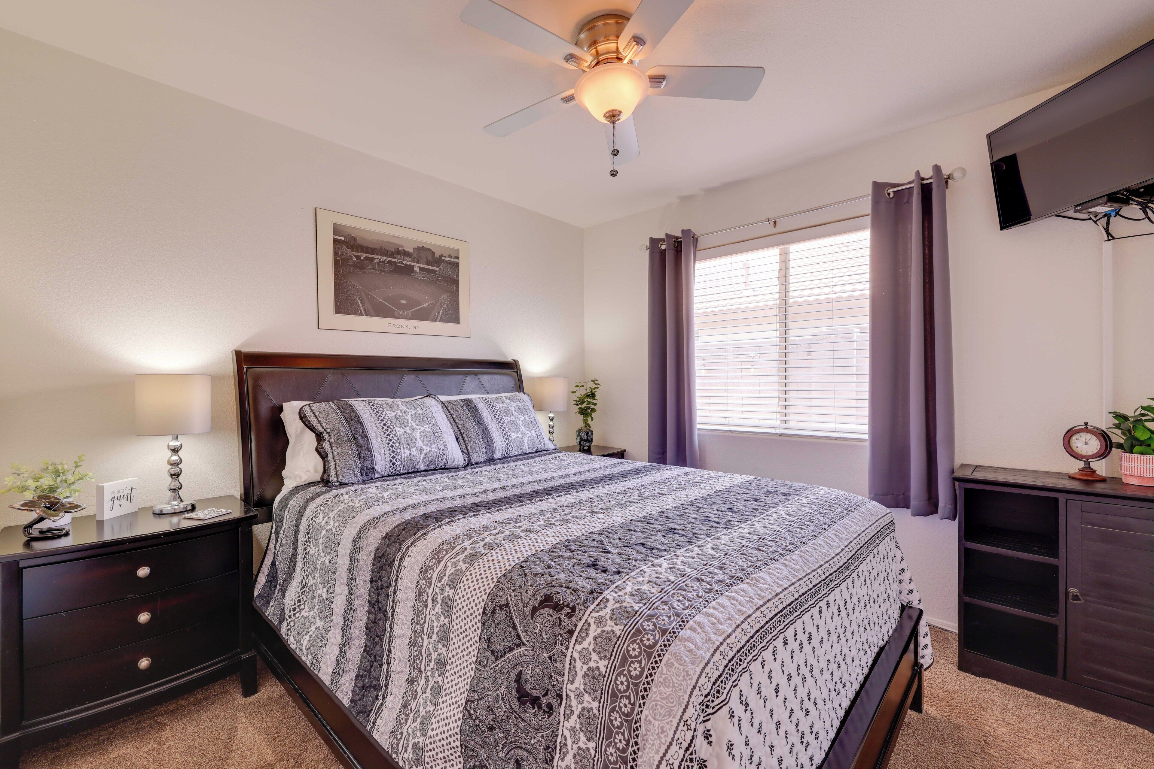 A bedroom with a bed, a nightstand, a ceiling fan, a window with curtains, a wall-mounted TV, and a framed picture.