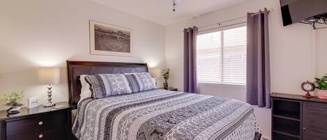 A bedroom with a bed, a nightstand, a ceiling fan, a window with curtains, a wall-mounted TV, and a framed picture.