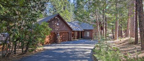 A cabin with a sloped roof, surrounded by tall pine trees, and a paved driveway leading to the entrance.