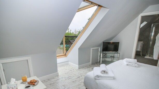 Une chambre moderne avec une lucarne, un lit, une table de chevet sur laquelle se trouvent un verre de jus d’orange et des croissants, ainsi qu’une télévision.