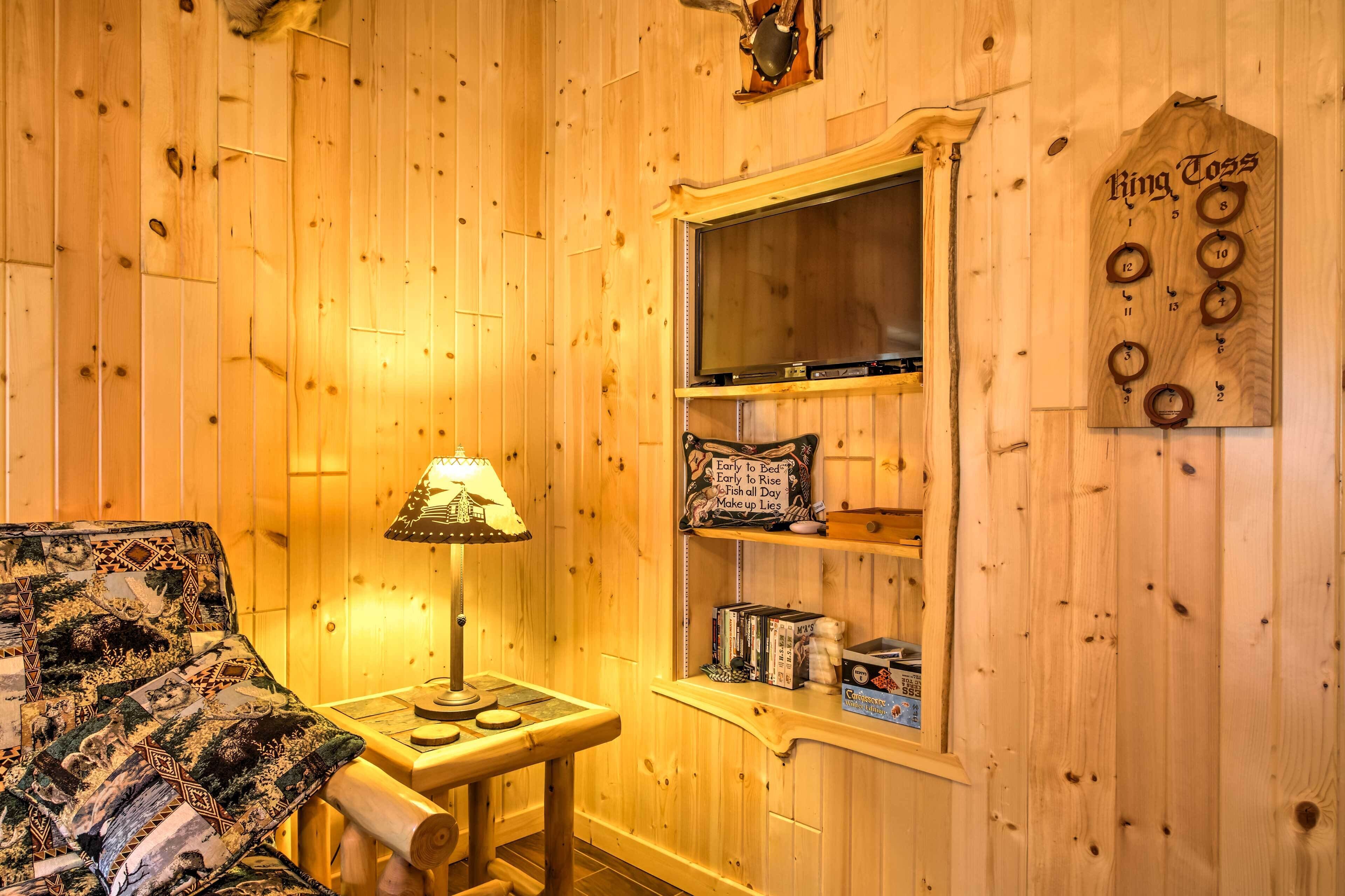 A cozy room with wooden walls, a bed with a patterned blanket, a small table with a lamp, and a shelf with books and a 'King Toss' sign.