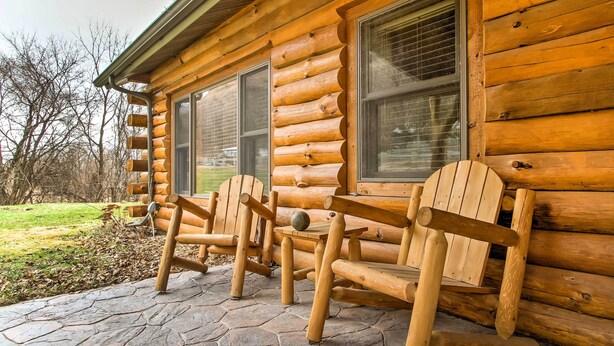 Een vakantiehuis in blokhutstijl met een terras van natuursteen, houten Adirondack-stoelen en een raam met luiken.