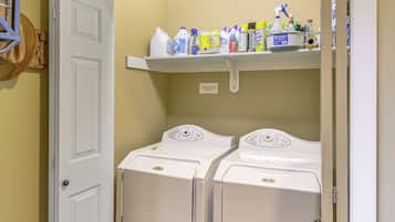 A laundry room with two front-loading washing machines and a shelf above containing various cleaning supplies.