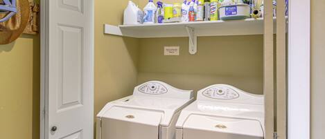 A laundry room with two front-loading washing machines and a shelf above containing various cleaning supplies.