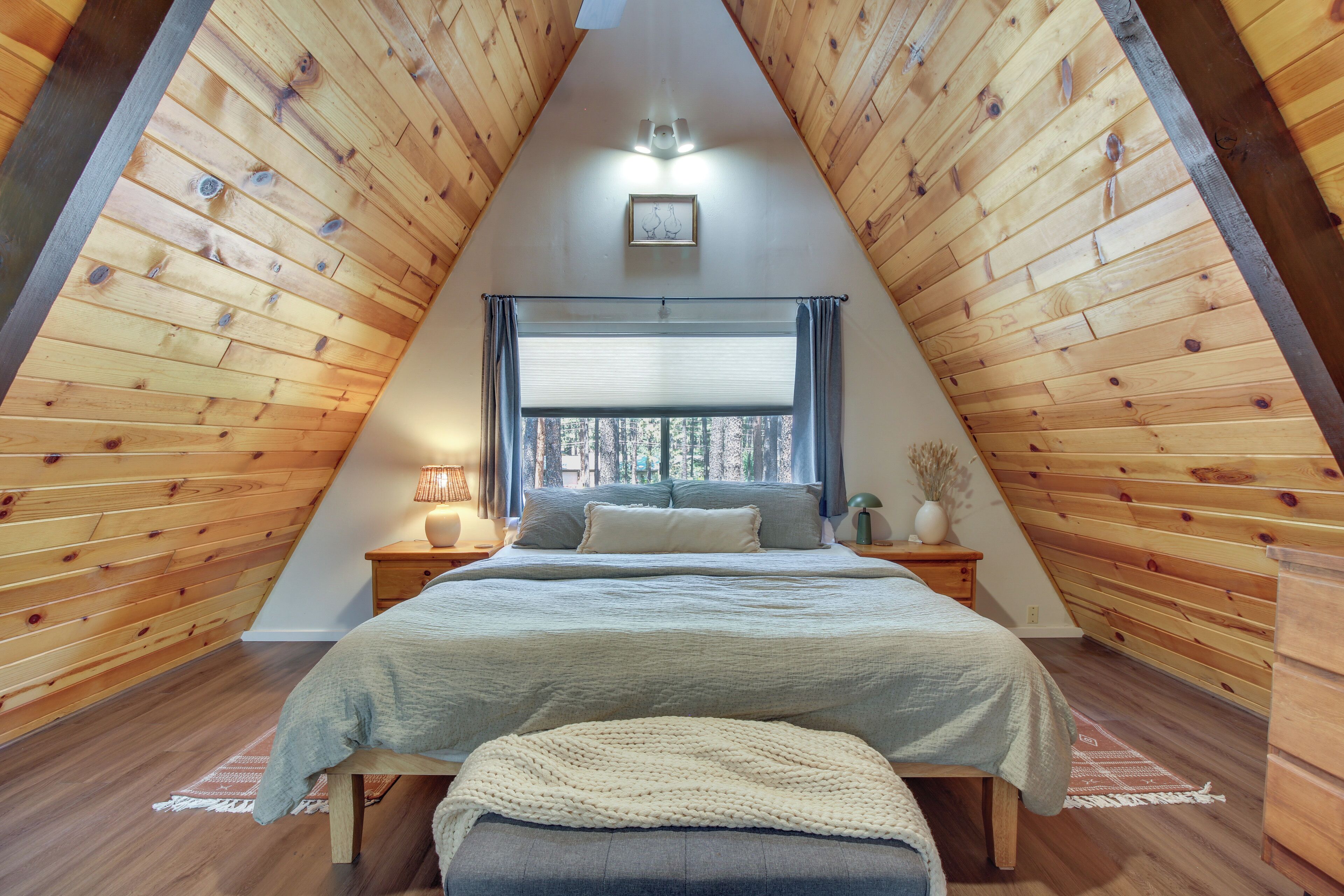 Una habitación triangular con paredes de madera, una cama con una manta gris, una mesita de noche con una lámpara y una ventana con cortinas.