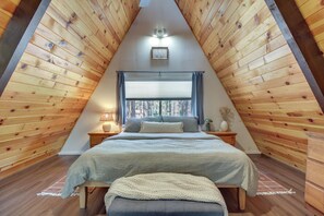 A triangular room with wooden walls, a bed with a grey blanket, a nightstand with a lamp, and a window with curtains. - The Silly Goose A-frame Cabin in Pinetop-lakeside (Pinetop)
