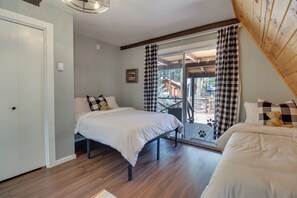 A bedroom with two beds, a window with checkered curtains, and a view of a wooden deck and trees.
