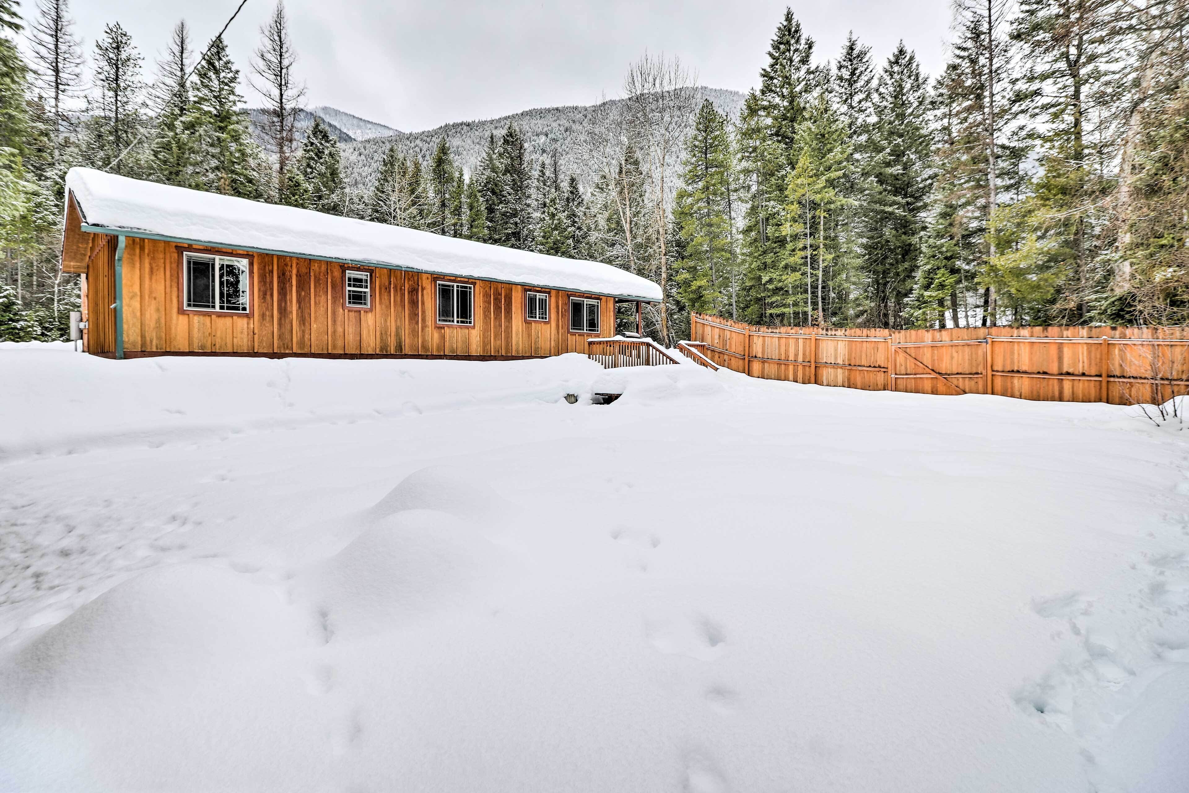 A wooden cabin with a snow-covered roof, surrounded by a wooden fence and tall evergreen trees.