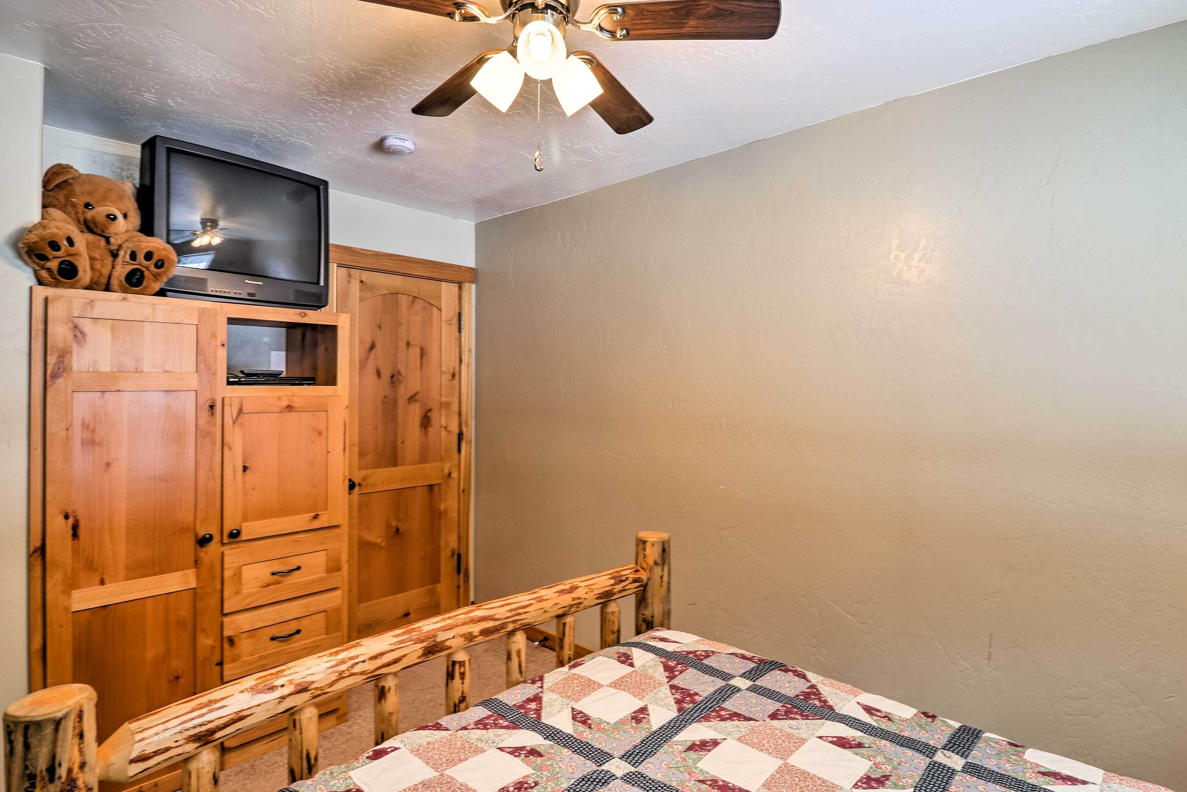 A bedroom with a wooden bed frame, a quilt, a wooden wardrobe, a television on top of the wardrobe, and a ceiling fan.