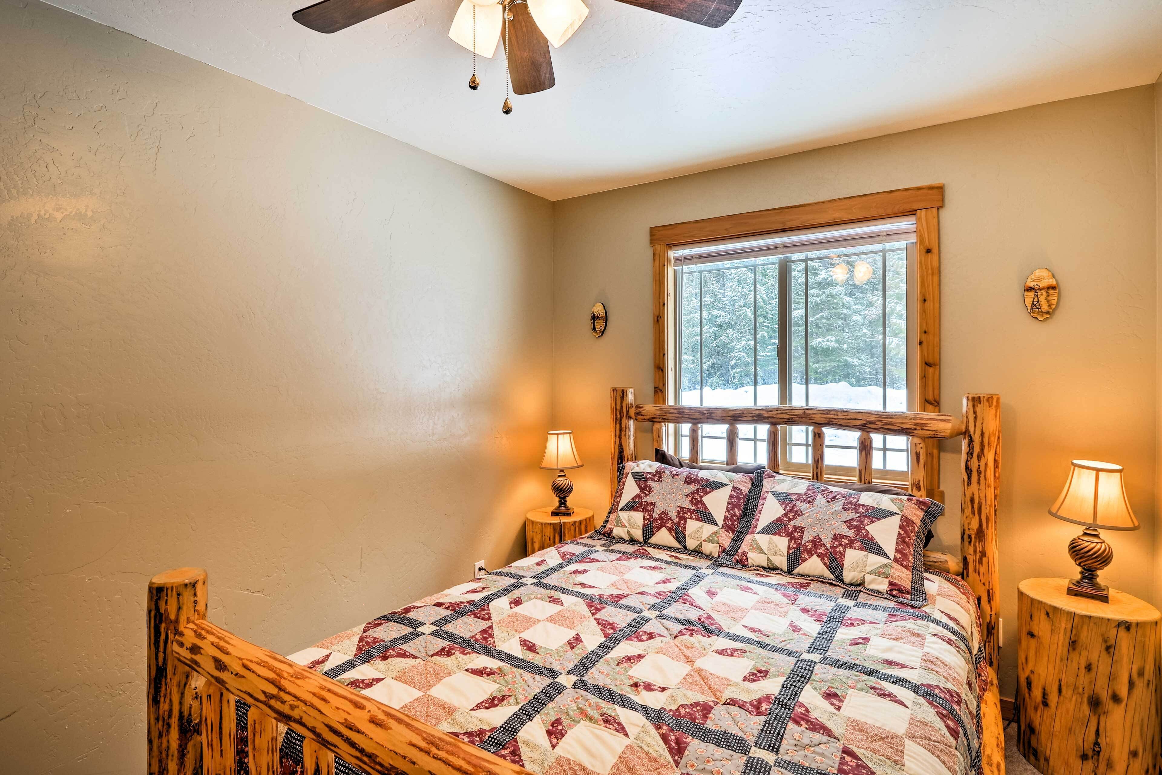 A cozy bedroom with a wooden bed frame, a quilted bedspread, two bedside lamps, a window with a view of trees, and a wooden ceiling fan.