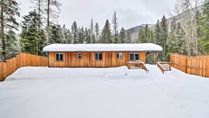 A wooden cabin with a snow-covered roof, surrounded by a wooden fence and snow-covered ground, with trees and mountains in the background. - Hungry Horse Cabin: 11 Mi to Glacier National Park (Hungry Horse)