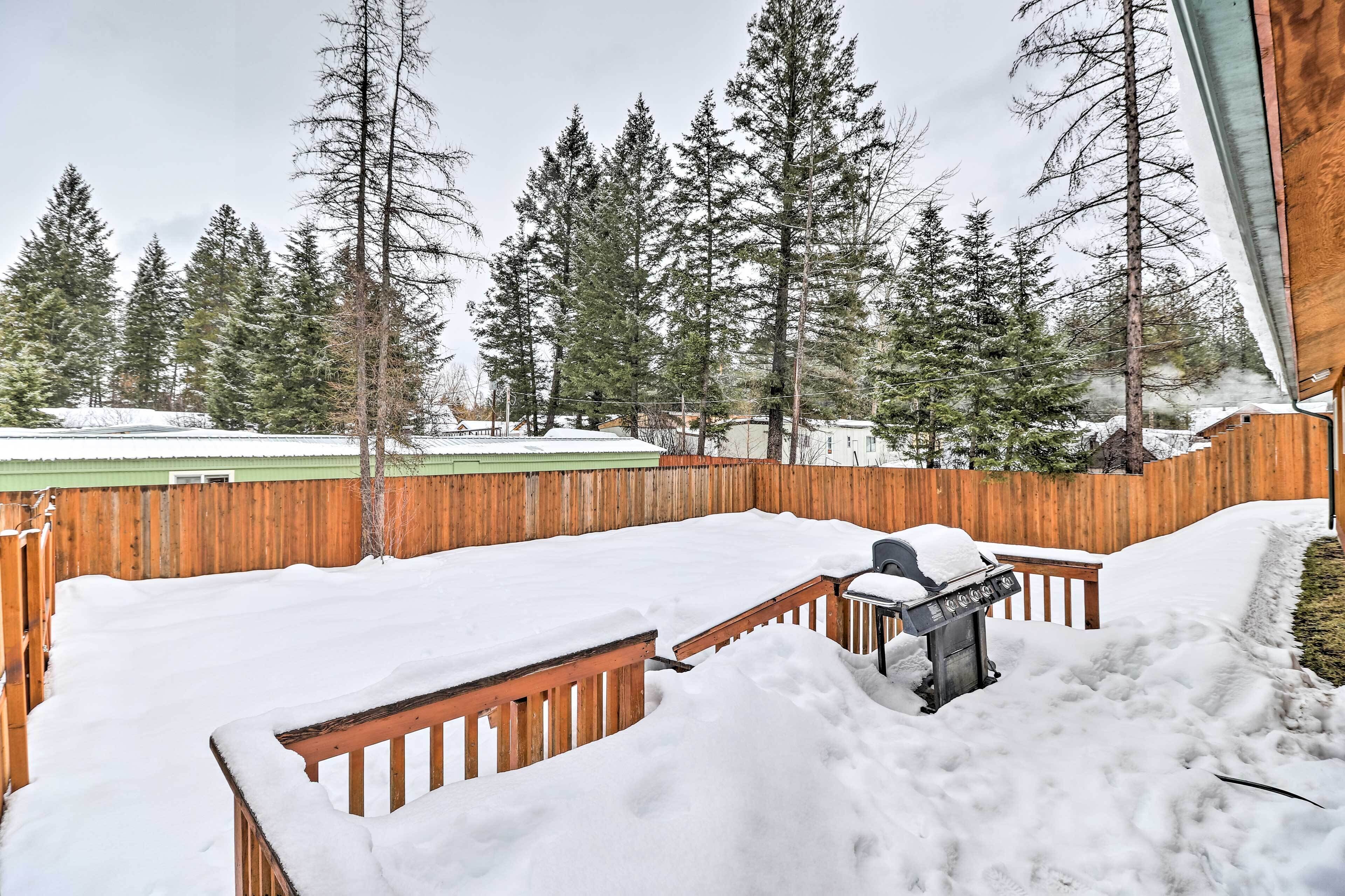 Un patio coperto di neve con una griglia per barbecue, circondato da una recinzione in legno e alti alberi sempreverdi.