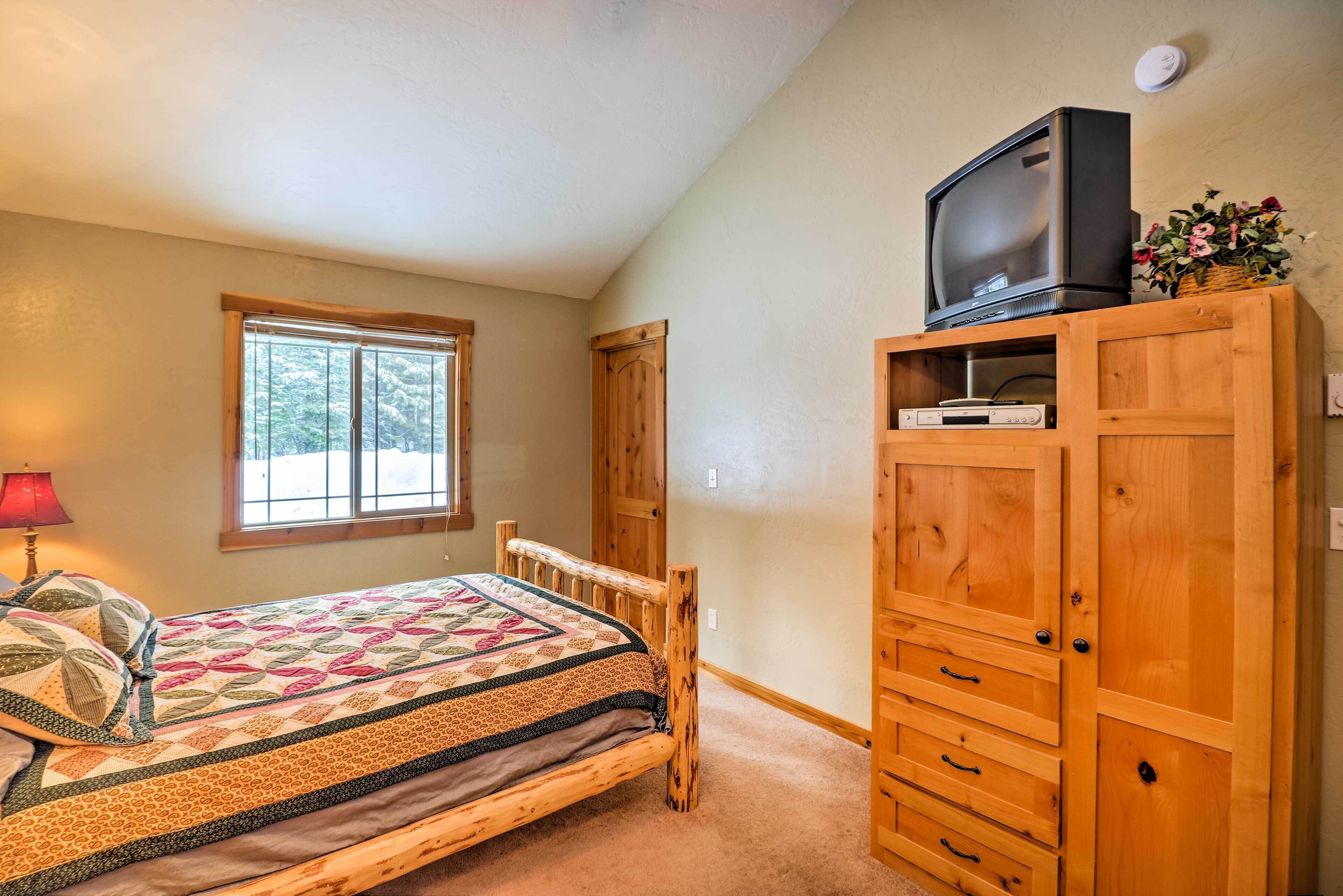 A bedroom with a wooden bed, a television on a wooden cabinet, a window with a view of trees, and a red lamp.
