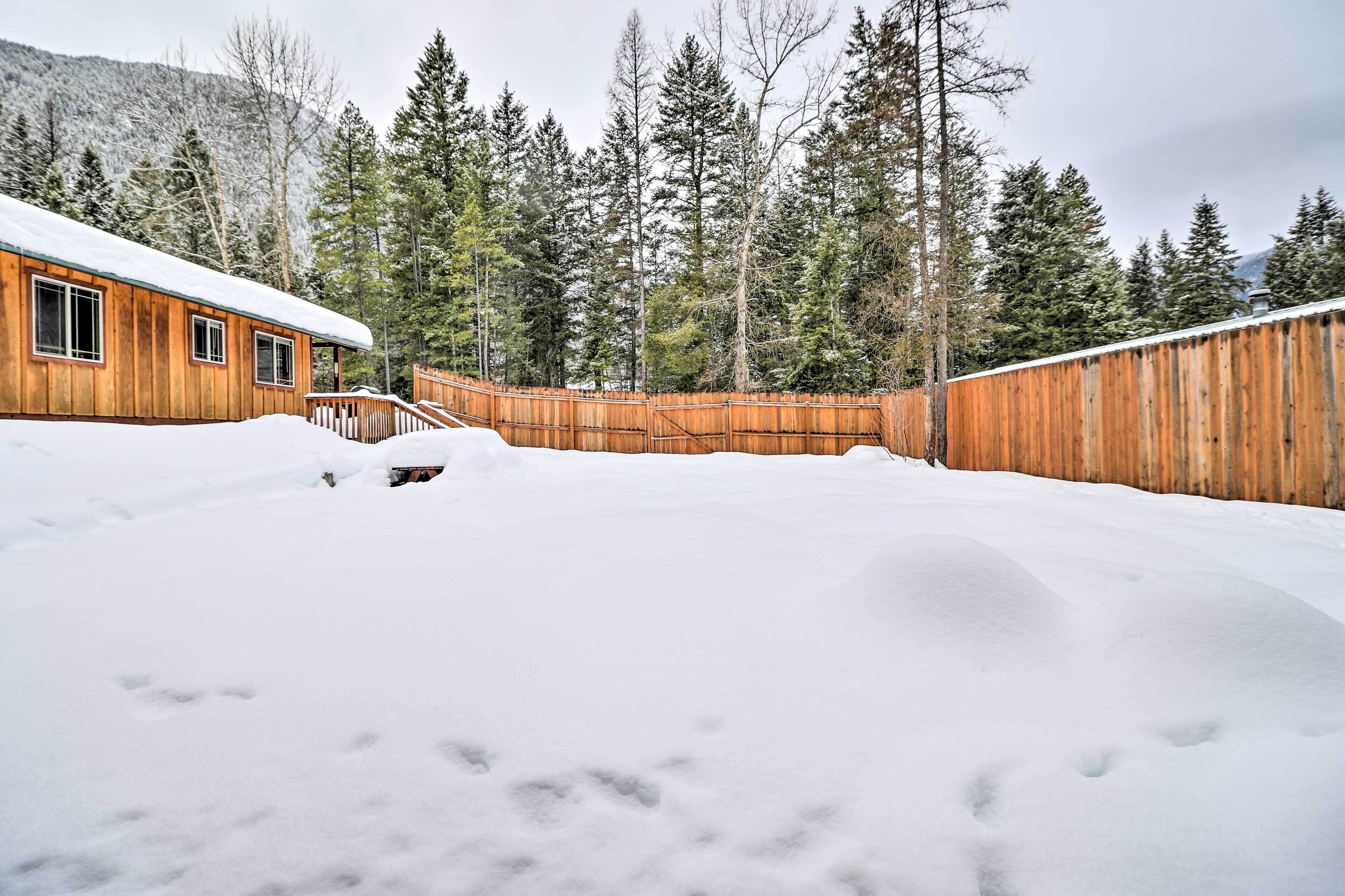 A wooden cabin with a snow-covered roof, surrounded by a wooden fence and dense evergreen trees.