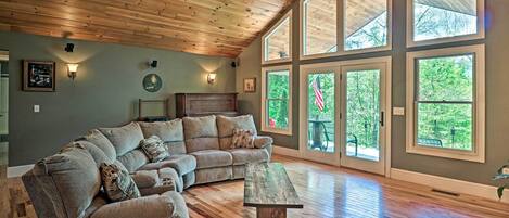 A spacious living room with a large sectional sofa, a coffee table, and a ceiling fan. Large windows provide natural light and views of the outdoors.