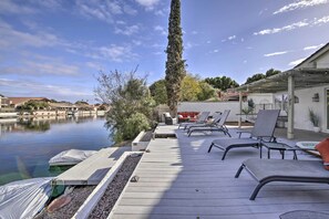A deck with lounge chairs overlooking a body of water, a tall tree, and a building with a covered patio.