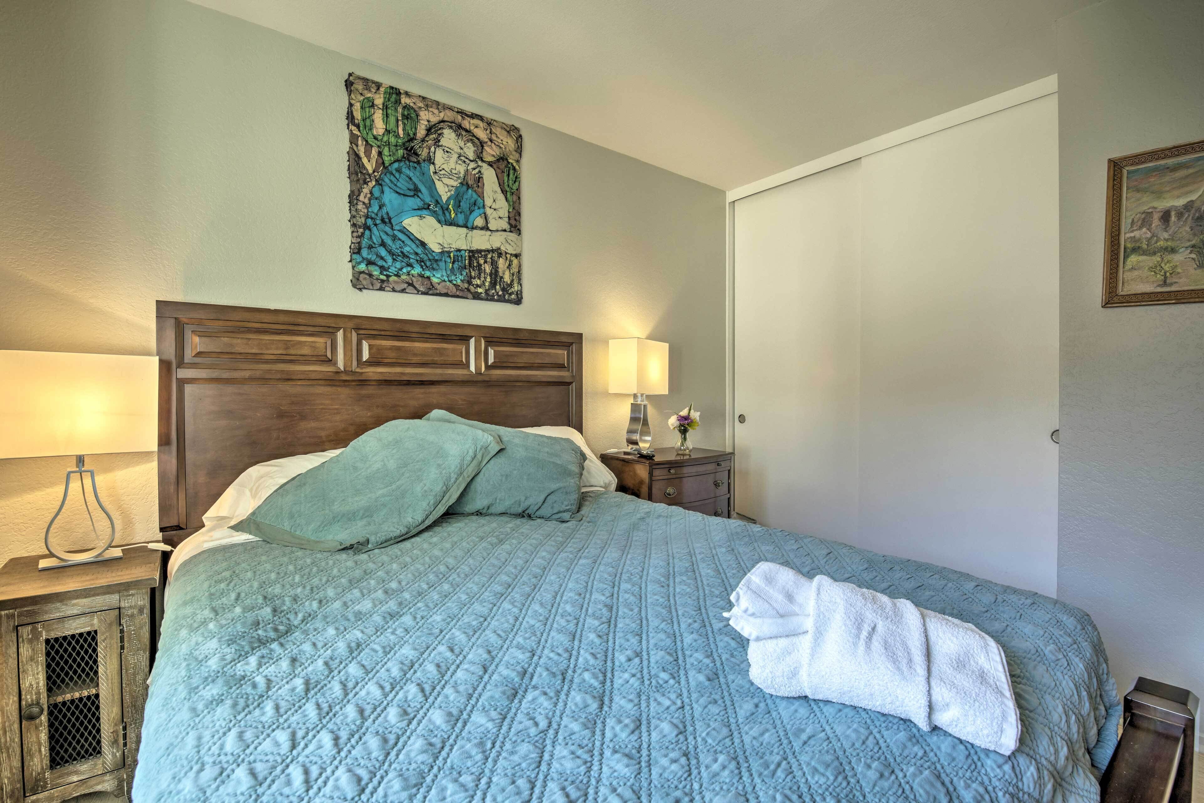 A neatly made bed with a blue quilt, a wooden headboard, a nightstand with a lamp, and a painting on the wall.