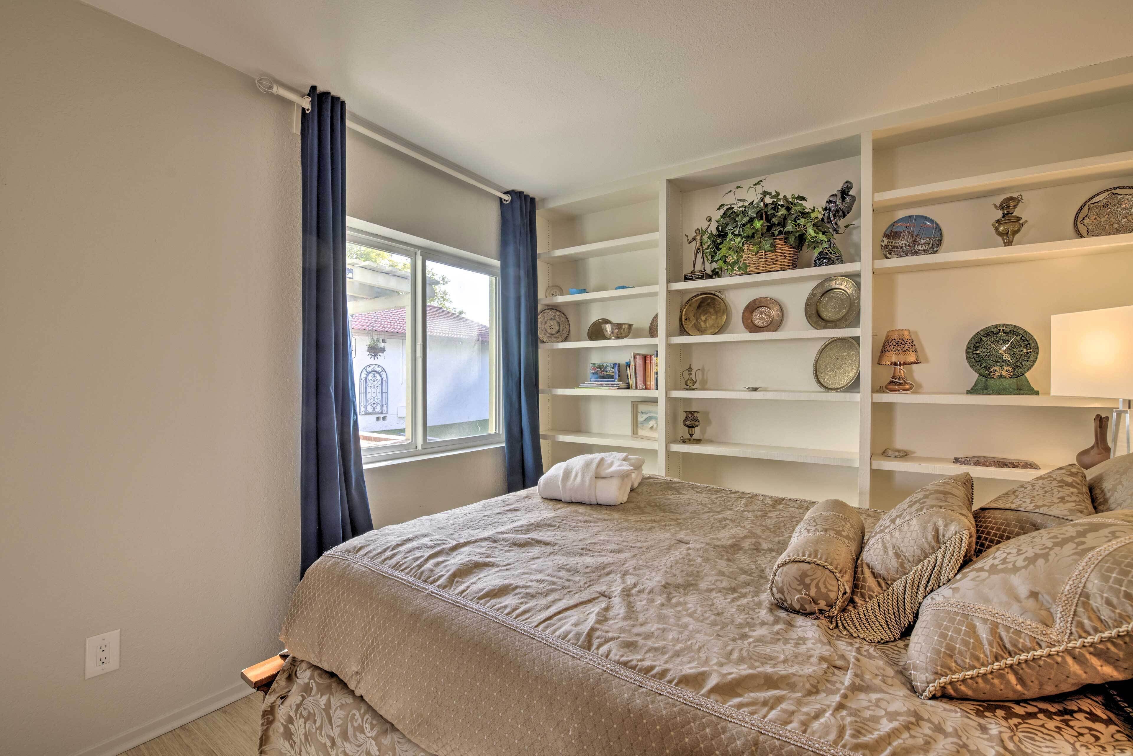 A bedroom with a large bed, a window with curtains, and a bookshelf with decorative items.