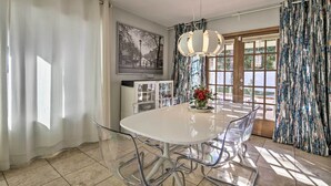 Un comedor con una mesa y sillas blancas, una araña y vistas a través de un ventanal.