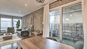 Ein modernes Wohnzimmer mit einem Holzesstisch, einem grünen Sessel und einer Couch. Eine Ziegelwand und ein großes Schiebeglasfenster führen in einen Außenbereich mit Stühlen und Pflanzen.