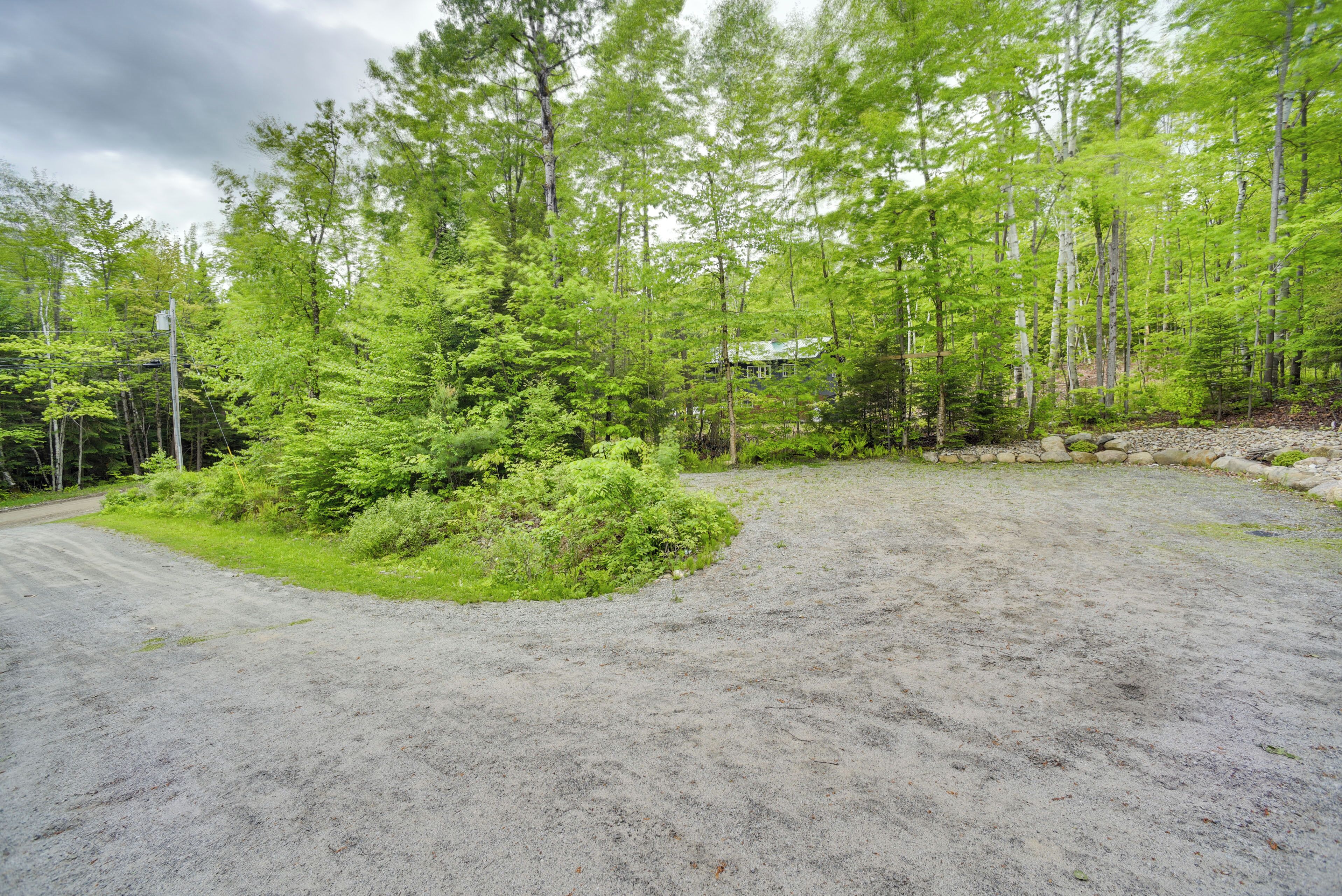 A gravel parking area with a dirt path leading to a house surrounded by dense green forest.