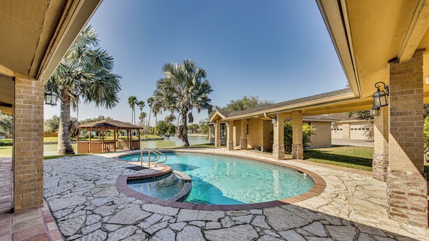 A swimming pool with a stone deck, surrounded by palm trees and a covered patio area. - Pool & Game Room: Waterfront Harlingen Home (Harlingen)
