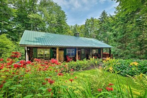 A rustic cabin with a green metal roof, surrounded by lush greenery and vibrant flowers. - Private Wooded Cabin: 7 Mi to Mount Snow! (Wilmington)